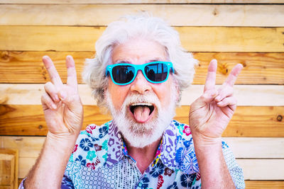 Portrait of happy senior man wearing sunglasses while showing victory sign against wooden wall