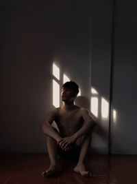 Young man looking away while sitting on floor against wall at home