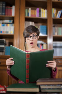 Portrait of surprised woman holding book against bookshelf in library