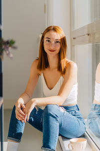 Smiling young red haired female in jeans and white top with bare shoulders sitting on floor near window and looking at camera in light room