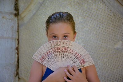 Portrait of girl holding folding fan while standing against wall
