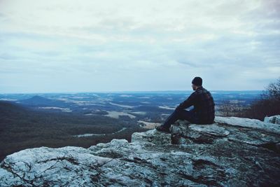 Man sitting on cliff while looking at landscape against sky