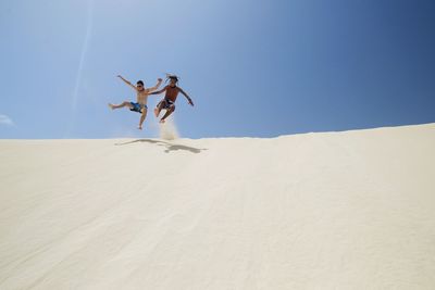 Low angle view of shirtless friends jumping on sand dune against blue sky