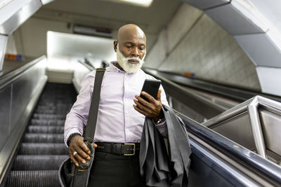 Senior commuter with laptop bag using smart phone on escalator