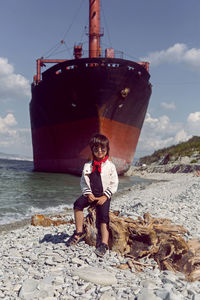 Fashionable boy child with long hair sits on a log next to a large ship that ran aground