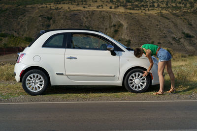 Young woman takes care of car checking rims on wheel against slopes in countryside on sunny day