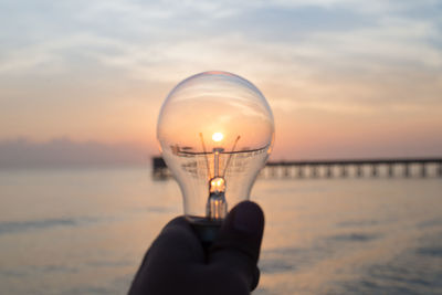 Person holding light bulb against sea during sunset