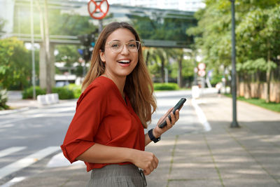Brazilian lively confident woman smiling at camera holding smartphone in sustainable metropolis