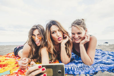 Happy friends taking selfie with smart phone while lying at beach