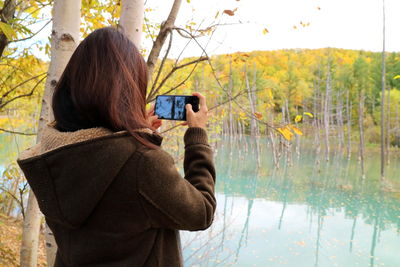 Back view of woman take a photo by mobile phone with blue pond in autumn of biei, hokkaido.