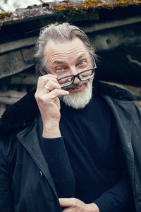 Elderly man with a beard stands in the village near a wooden shed in a sheepskin coat and glasses