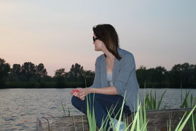Young woman sitting by lake against clear sky during sunset