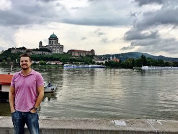 Man with hands in pockets standing by lake against cloudy sky at esztergom basilica