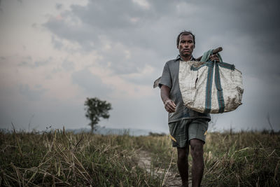 Portrait of man carrying bag while walking on field against sky