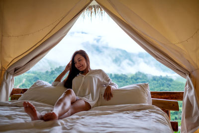 A young woman lying down on a white bed in the morning with a beautiful nature view outside the tent