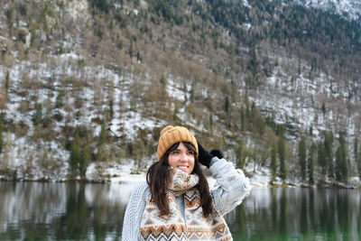 Young woman in winter sweater smiling, standing by a lake.