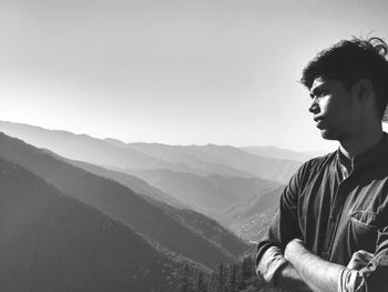 Thoughtful young man with arms crossed standing on mountain against clear sky during sunny day