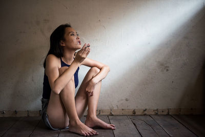 Full length of young woman smoking cigarette while sitting on floor against wall at home