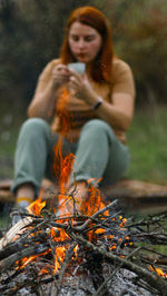 Woman drinking coffee while sitting against campfire