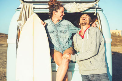 Couple with surfboard and caravan laughing on field