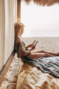 Side view of woman reading book while sitting in cabana on vacation at beach