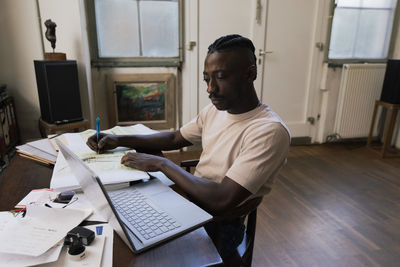 Male entrepreneur using laptop and writing on document at home office