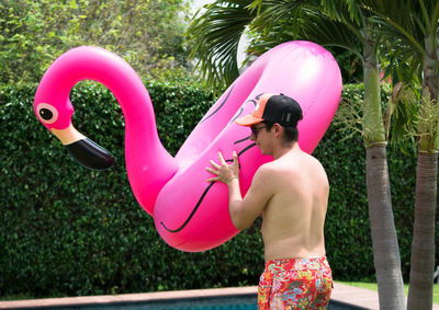 Shirtless young man holding pink flamingo shape inflatable ring at poolside