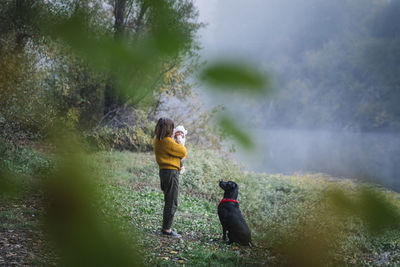 A woman is holding a baby and looking at a dog near a river