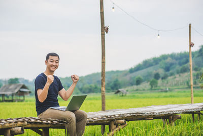 Portrait of man using laptop while sitting on boardwalk at rice paddy