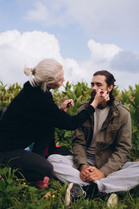 Woman doing make-up on male friend sitting on grass against sky