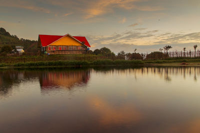 House by lake and buildings against sky during sunset