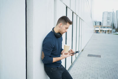 Side view of a modern man in casual clothes sitting near a white wall