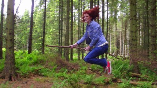 Full length of woman jumping while sitting on stick at forest