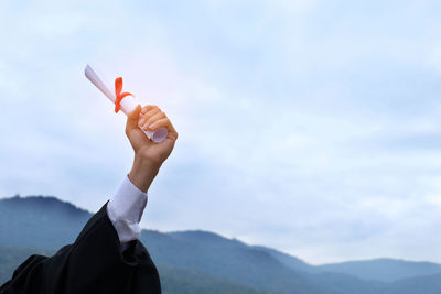 Cropped hand of woman holding diploma against sky