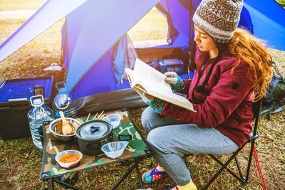 Young woman reading book while sitting on chair at campsite