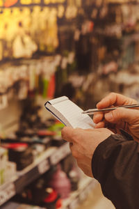 Female hands holding paper notebook of shopping list, blurred shelves on background. partial view of