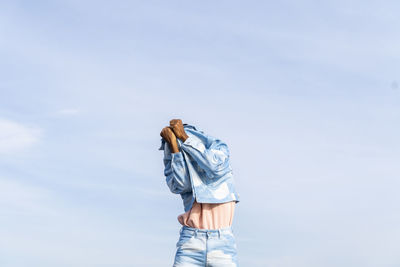 Man hiding face with jacket while standing against sky