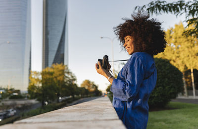 Smiling woman photographing through camera by wall