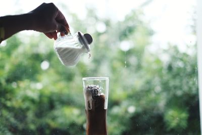 Cropped hand pouring milk into drinking glass against window at home
