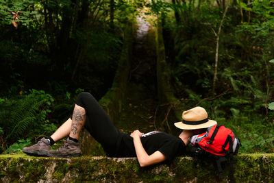 Side view of unrecognizable female explorer lying on backpack in dark woods and sleeping while resting during trekking