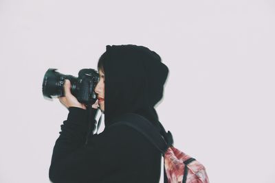 Side view of young woman photographing while standing against white background