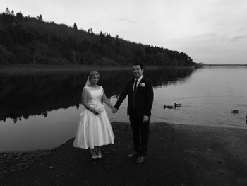 Portrait of wedding couple holding hands while standing at lakeshore against sky