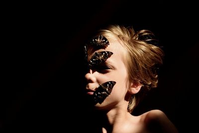 Close-up of boy with butterflies on face against black background