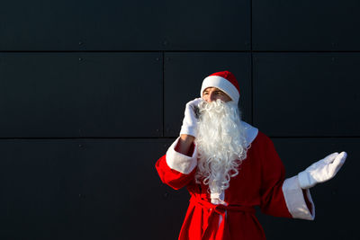 Man wearing santa clause costume talking on phone while standing against wall outdoors