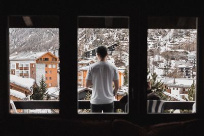 Rear view of man standing on balcony seen through window during winter