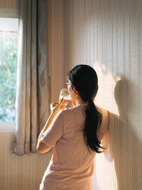 Rear view of woman drinking coffee while standing by wall at home