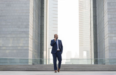 Full length of businessman talking on mobile phone while walking against office buildings