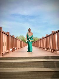 Full length portrait of woman standing on footbridge against sky