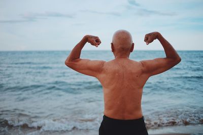 Rear view of shirtless senior man flexing muscles while standing at beach against sky during sunset