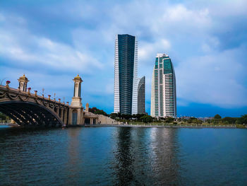 View of modern buildings by river against cloudy sky